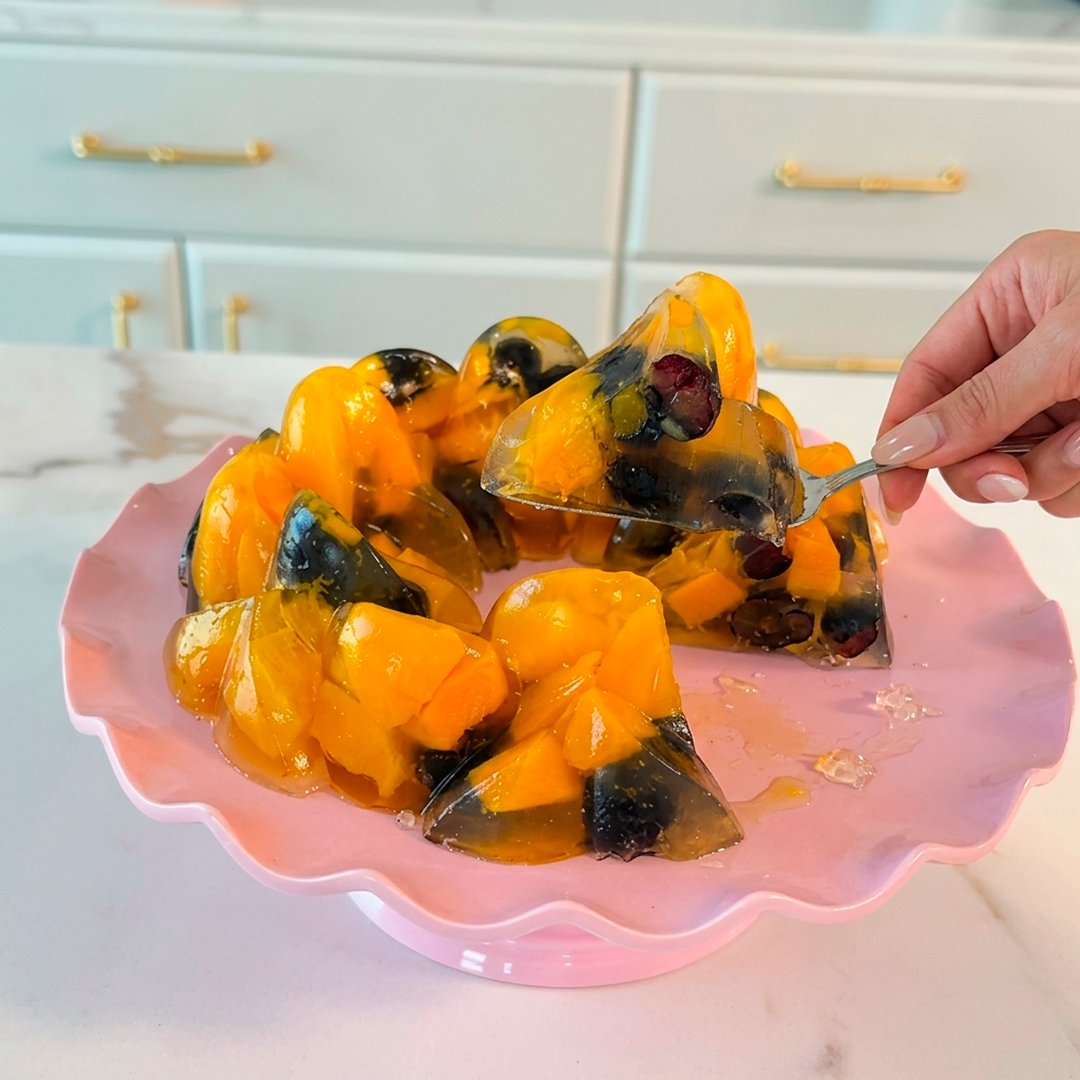 Mango and blueberry fruit jelly ring unmoulded on a pink cake stand, hand lifting a slice with a cake server, kitchen background